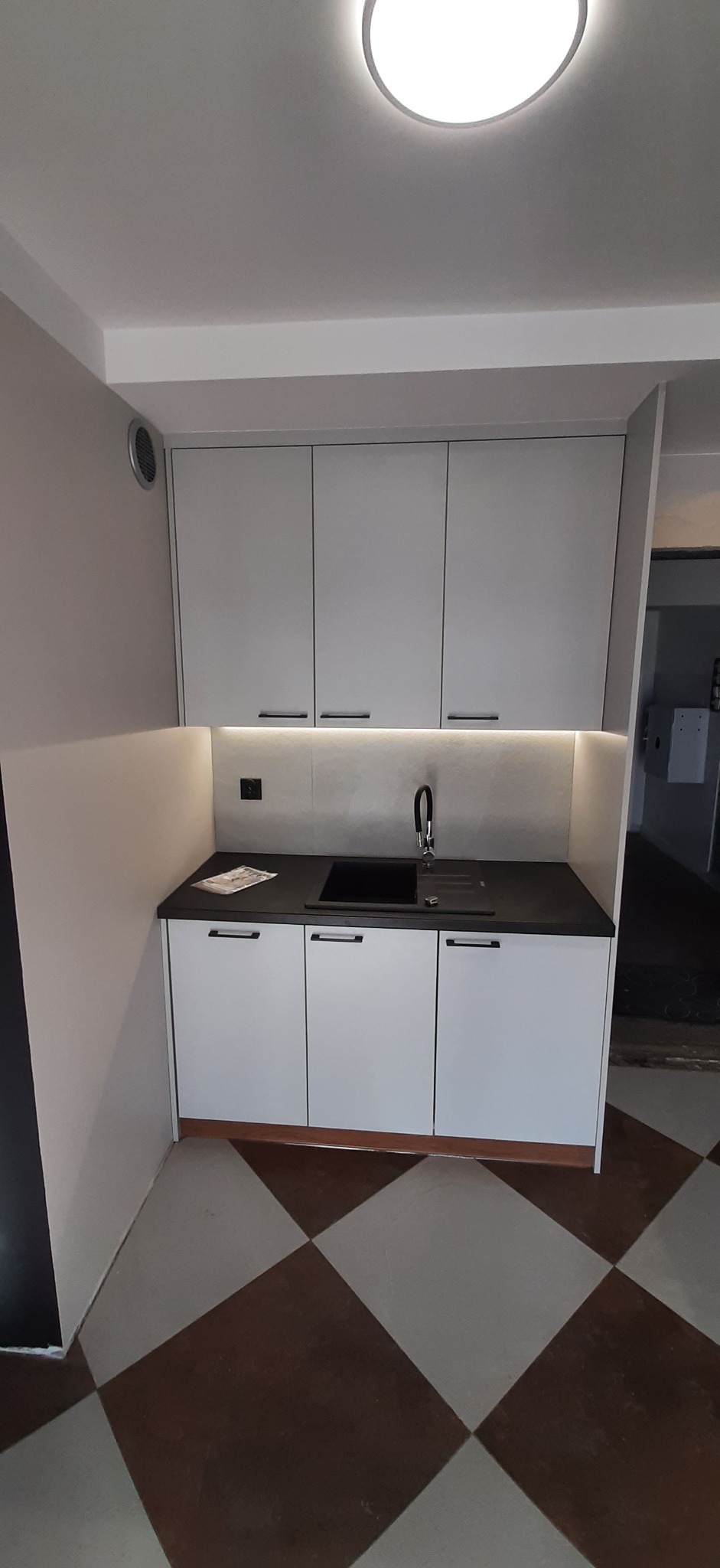 Nowoczesna, minimalistyczna kuchnia z białymi szafkami, czarnym blatem i podświetleniem LED. Uwagę przykuwa podłoga w brązowo-beżową szachownicę i nietypowe wykończenie ściany.