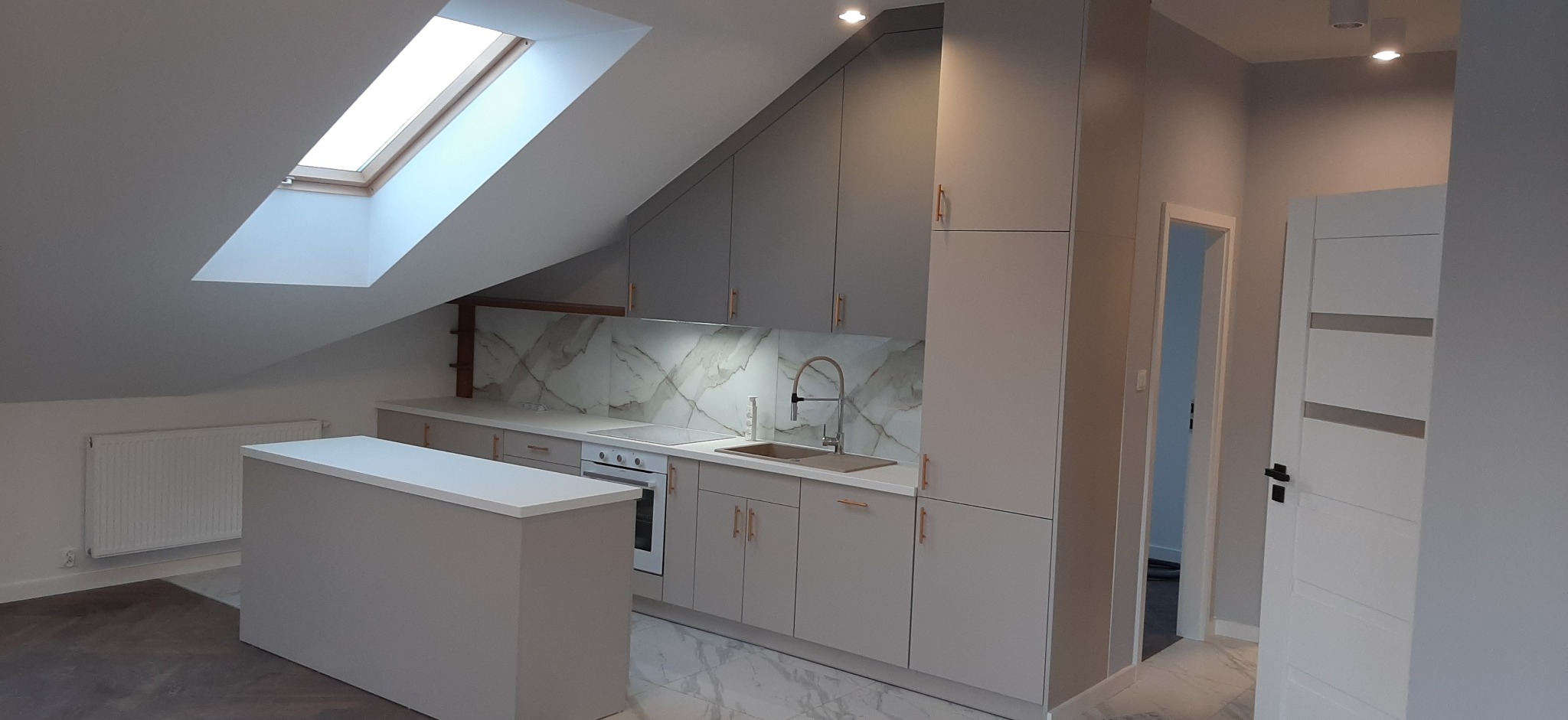 Jasnoszara kuchnia na poddaszu ze skośnym sufitem i oknem dachowym. Marmurowy backsplash i złote uchwyty dodają elegancji. Wyspa kuchenna z białym blatem.