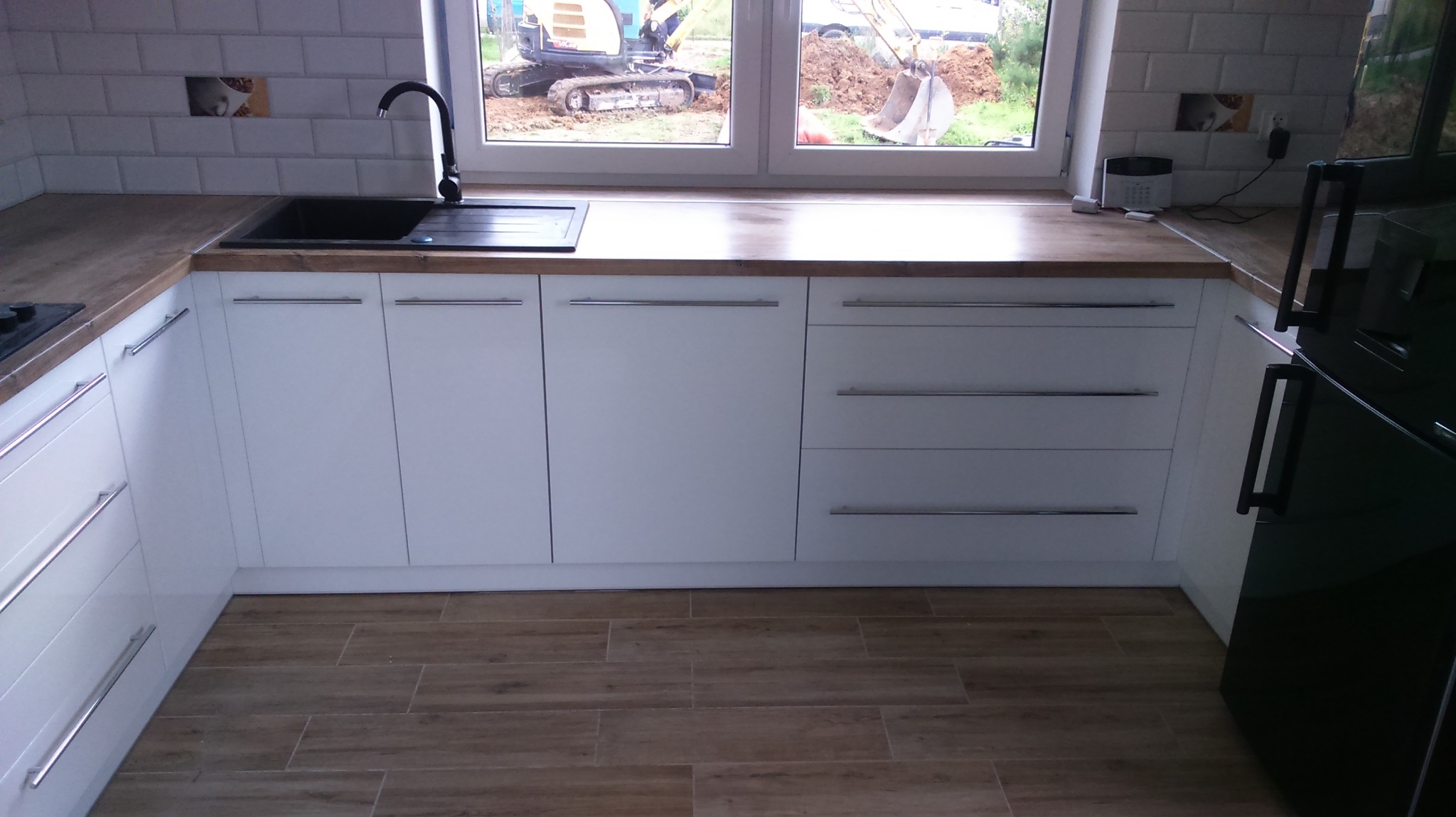 Biała zabudowa kuchenna z drewnianym blatem i czarnym zlewem, widok na okno z koparką na zewnątrz. Minimalistyczny design, płytki imitujące drewno na podłodze.
