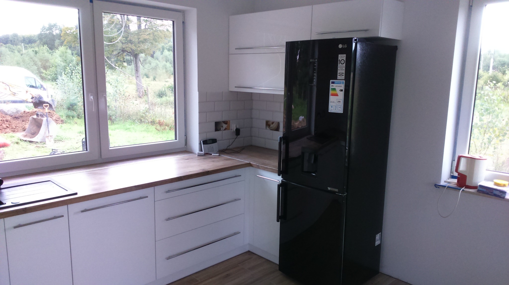 Nowoczesna kuchnia: białe szafki z drewnianym blatem, czarna lodówka LG, okno z widokiem na zewnątrz. Uwagę zwraca subtelne oświetlenie i minimalistyczny design.