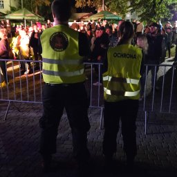 AGENCJA OCHRONY OSÓB I MIENIA COBRA 9 - Dwóch ochroniarzy w żółtych kamizelkach stoi tyłem do kamery, oddzielając metalowym ogrodzeniem tłum ludzi podczas nocnego wydarzenia plenerowego.