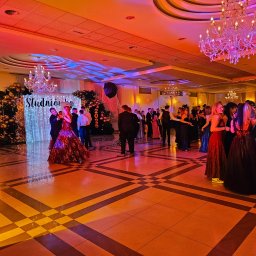 AGENCJA OCHRONY OSÓB I MIENIA COBRA 9 - Sala balowa z tańczącymi ludźmi podczas studniówki 2026, dekoracje balonowe, kryształowe żyrandole, parkiet w szachownicę, oświetlenie sceniczne.