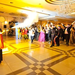 AGENCJA OCHRONY OSÓB I MIENIA COBRA 9 - Sala balowa w ciepłych barwach, goście z podniesionymi świetlnymi pałeczkami, kobieta obsługuje wyrzutnię dymu. Dekoracje balonowe w tle.