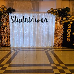 AGENCJA OCHRONY OSÓB I MIENIA COBRA 9 - Dekoracja studniówkowa z błyszczącą ścianką, białymi zasłonami z lampkami i girlandami z czarno-złotych balonów. Napis 'Studniówka' i rok 2026.