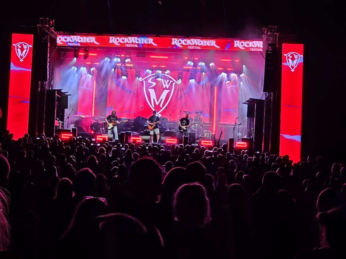 Tłum widzów na koncercie Rockwater Festival, scena oświetlona czerwonymi światłami, zespół na scenie gra muzykę. Ochrona imprez masowych.