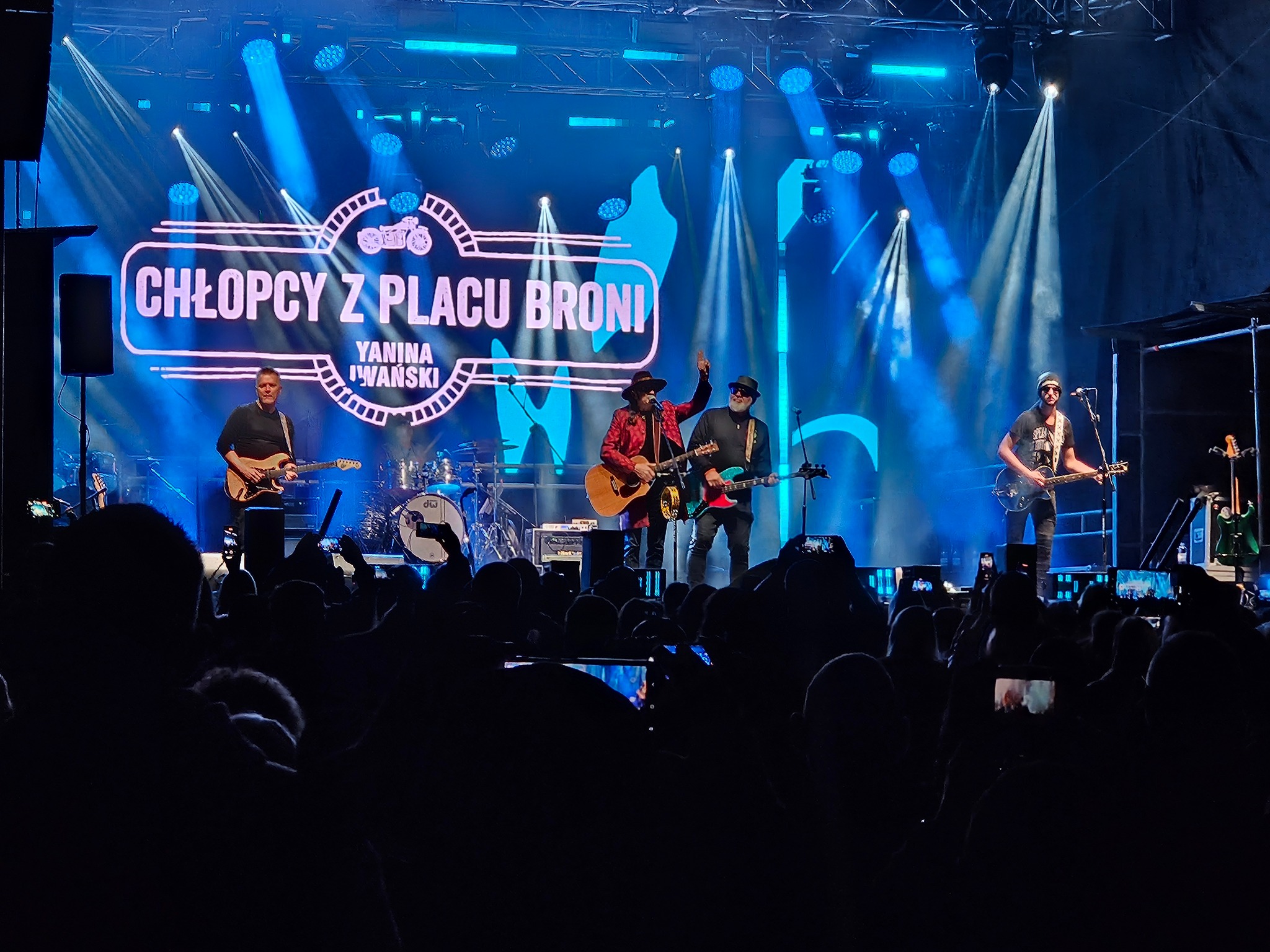 Zespół muzyczny 'Chłopcy z Placu Broni' na scenie podczas koncertu, oświetlony niebieskimi światłami, widownia nagrywa występ telefonami.