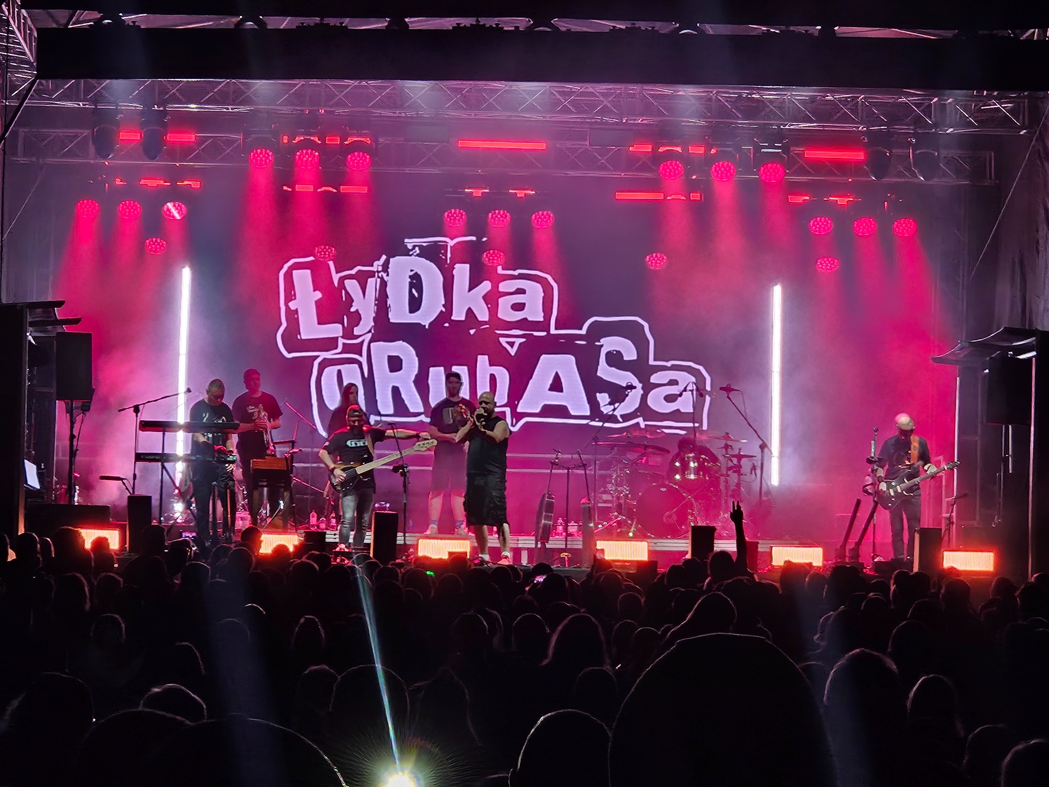 Zespół muzyczny Łydka Grubasa na scenie podczas koncertu. Czerwone światła reflektorów oświetlają muzyków i publiczność. Widoczny tłum fanów.
