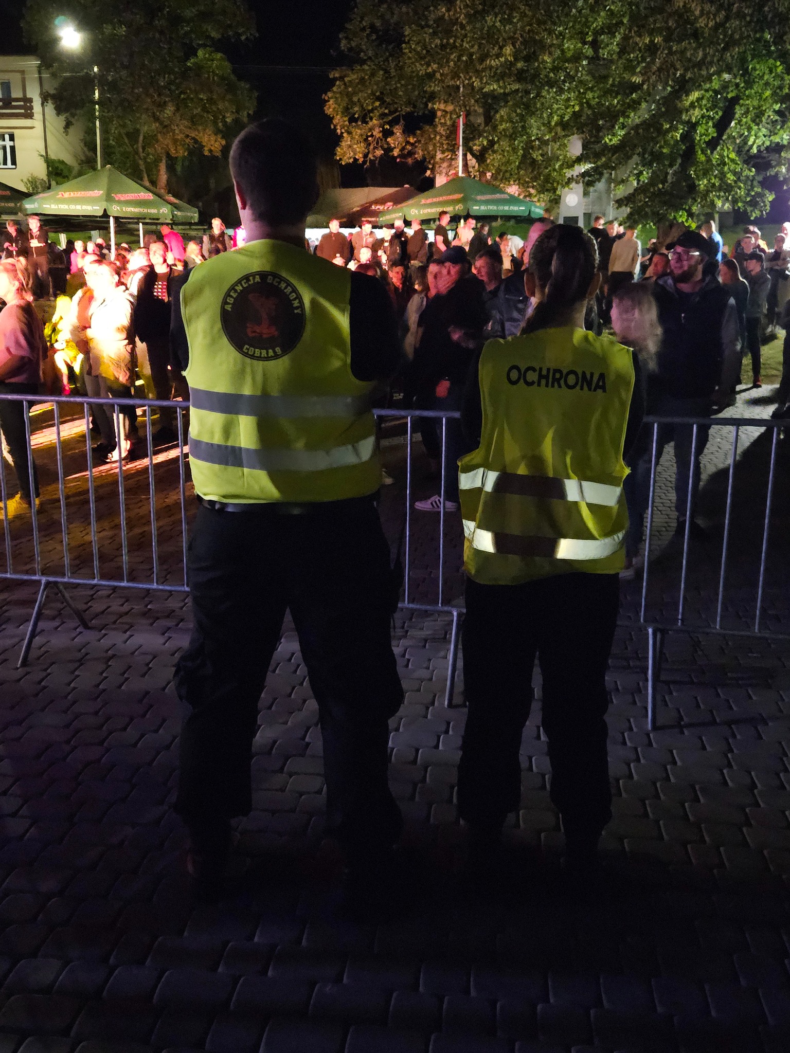 Dwóch ochroniarzy w żółtych kamizelkach stoi tyłem do kamery, oddzielając metalowym ogrodzeniem tłum ludzi podczas nocnego wydarzenia plenerowego.