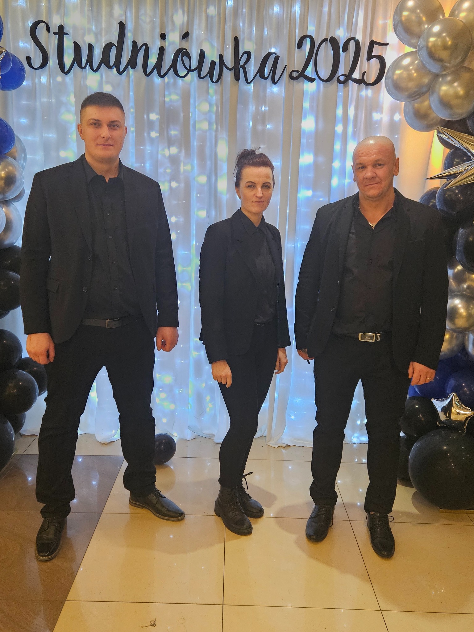 Trzech ochroniarzy w czarnych garniturach stoi na tle dekoracji studniówkowej z balonami i napisem 'Studniówka 2025'.
