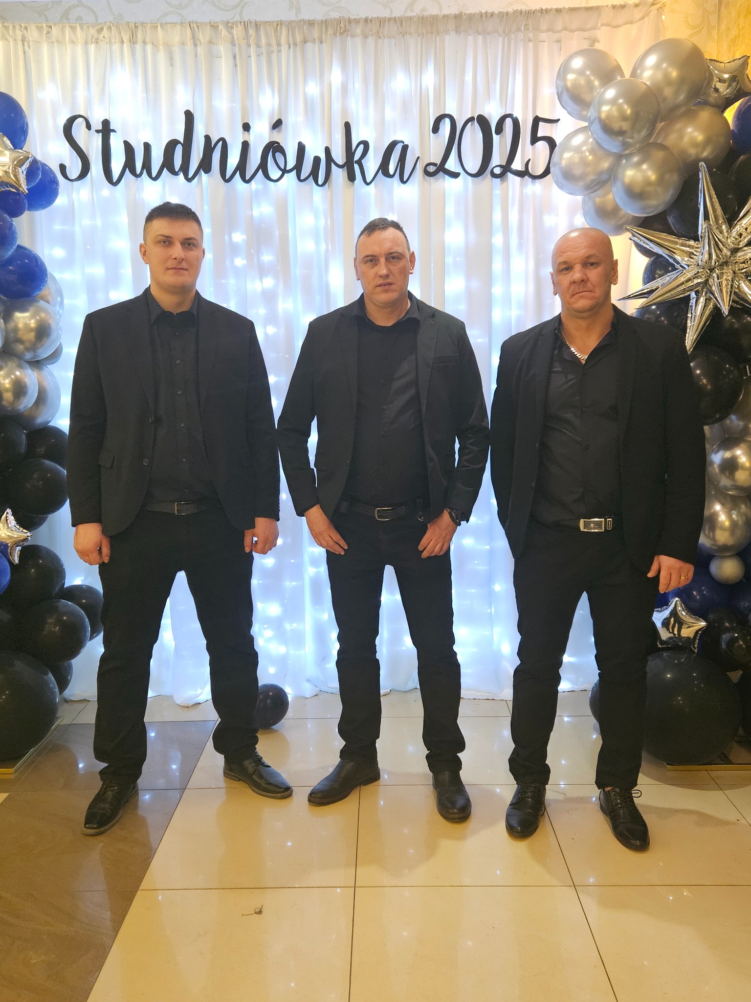 Trzech mężczyzn w czarnych garniturach pozuje na tle dekoracji studniówkowej z balonami i napisem 'Studniówka 2025'.