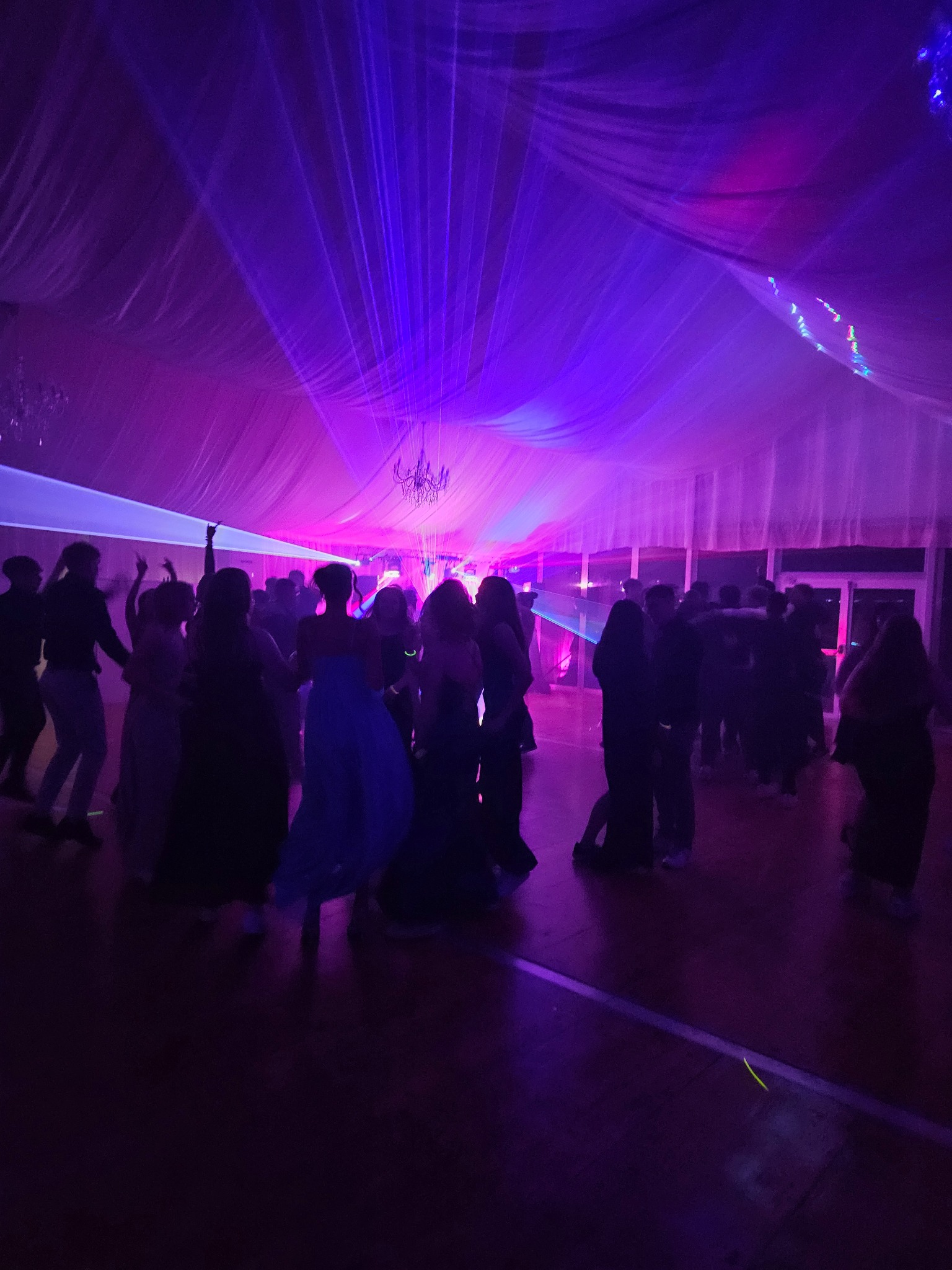 Zabawa w fioletach: Grupa osób tańczy w oświetlonym pomieszczeniu z dekoracyjnym sufitem i żyrandolem, tworząc dynamiczną scenę imprezową.