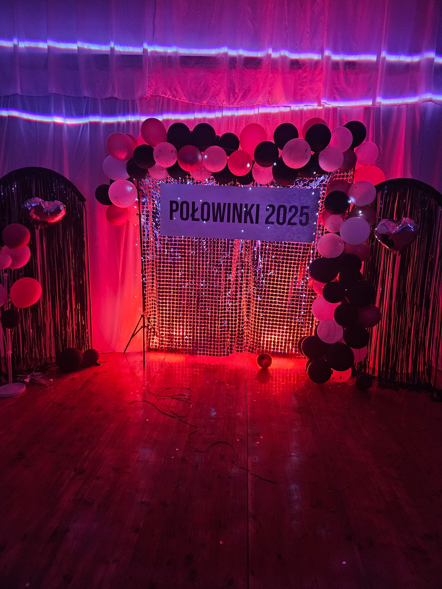 Dekoracja sali na połowinki 2025 z błyszczącym tłem, balonami w kolorach czerni, bieli i różu, oświetlona czerwonym światłem. Widoczne kurtyny i drewniana podłoga.