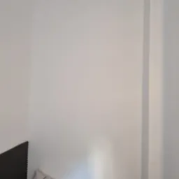 Biała ściana w rogu pokoju z widoczną listwą przypodłogową i fragmentem zagłówka łóżka. Świeżo pomalowana powierzchnia.