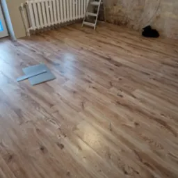 Ukończona podłoga z paneli drewnopodobnych w pokoju z grzejnikiem i drabiną. Dwa panele leżące luzem na podłodze. Widoczne ślady po rysunku na ścianie.