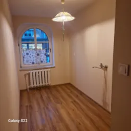 Pusta, jasna kuchnia po remoncie z panelami podłogowymi, grzejnikiem pod oknem i kranem na ścianie. Widok na ulicę przez okno z firanką.