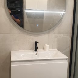 IVAN BILOZERSKYKH - Biała szafka pod umywalkę z czarną armaturą i okrągłym lustrem z podświetleniem LED w nowoczesnej łazience z szarymi płytkami.