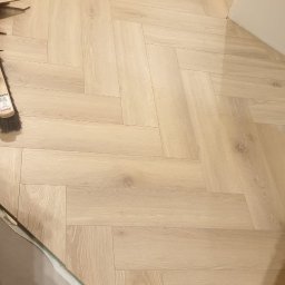 Good joob - Jasne panele ułożone w jodełkę, widoczne przycięte krawędzie przy ścianie i szczotka do zamiatania. Układanie paneli podłogowych w trakcie wykańczania.