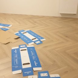 Good joob - Ułożone panele podłogowe w jodełkę, z rozłożonymi kartonami po panelach. Widok na jasną podłogę i fragment ściany z grzejnikiem.