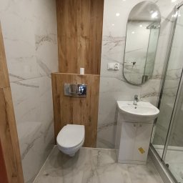 IVAN BILOZERSKYKH - Nowoczesna łazienka po remoncie: białe marmurowe płytki, drewniane akcenty, owalne lustro, biała umywalka z szafką i toaleta. Minimalistyczny design, jasne wnętrze.