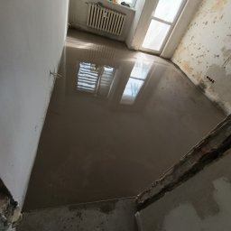 Good joob - Wylany, mokry podkład podłogowy w pokoju z oknem i grzejnikiem. Ściany częściowo przygotowane do malowania, widoczne ślady po usuniętych płytkach.
