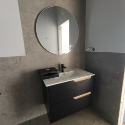 Good joob - Nowoczesna łazienka z okrągłym lustrem nad czarną szafką z umywalką. Szare płytki na ścianach i podłodze. Minimalistyczny design, widoczne niedokończone elementy.