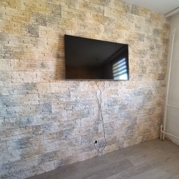 Good joob - Ściana wykończona dekoracyjną okładziną kamienną w odcieniach beżu i szarości. Na ścianie zawieszony czarny telewizor. Widoczny fragment podłogi z paneli.