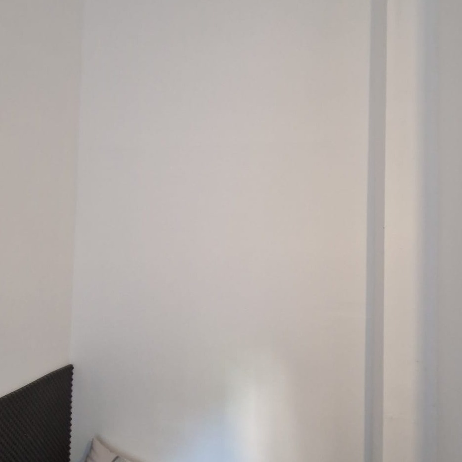 Biała ściana w rogu pokoju z widoczną listwą przypodłogową i fragmentem zagłówka łóżka. Świeżo pomalowana powierzchnia.