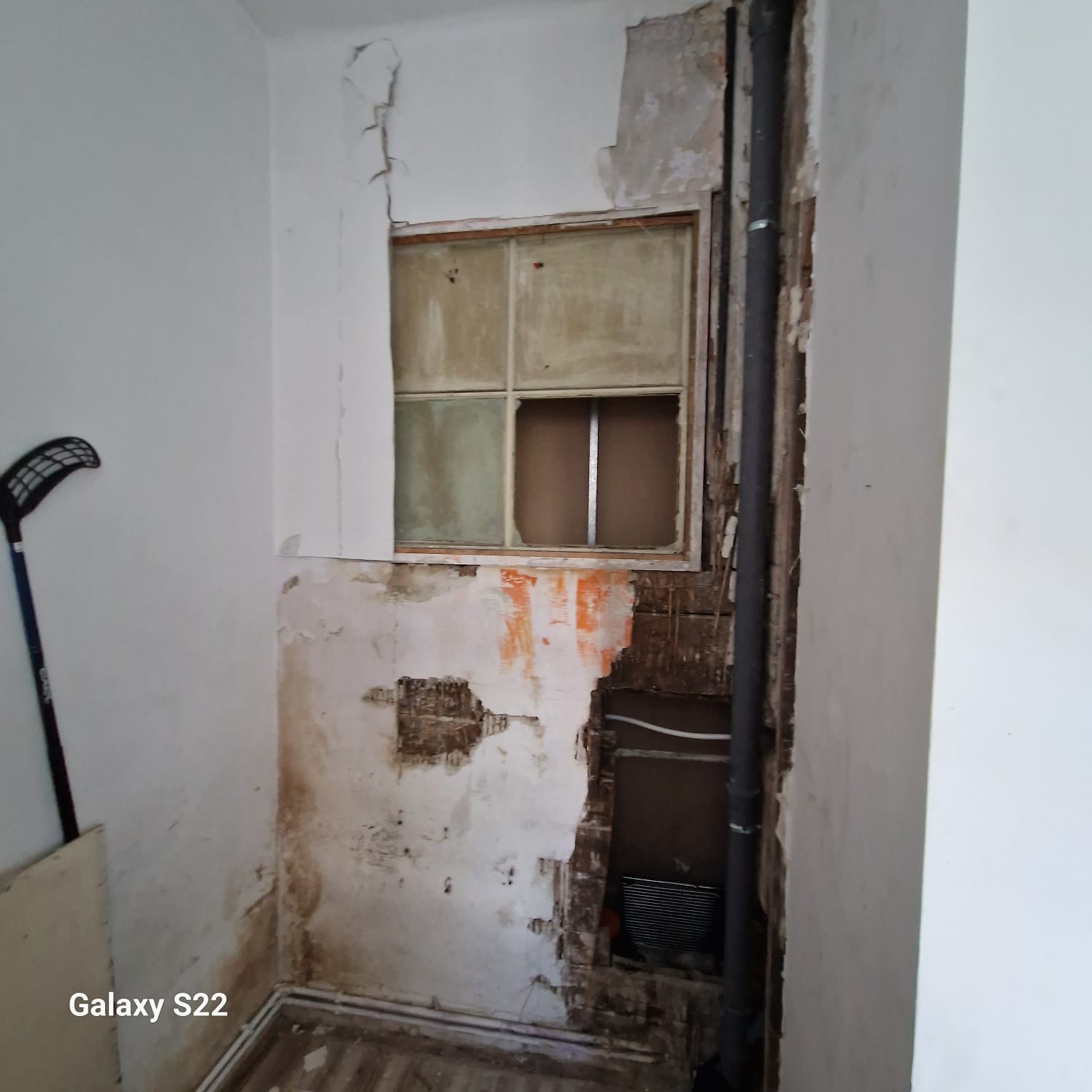 Wnętrze w trakcie demontażu: odsłonięte rury, fragmenty starej instalacji, uszkodzone ściany z resztkami tapet i farb. Widoczne okno i fragment podłogi.