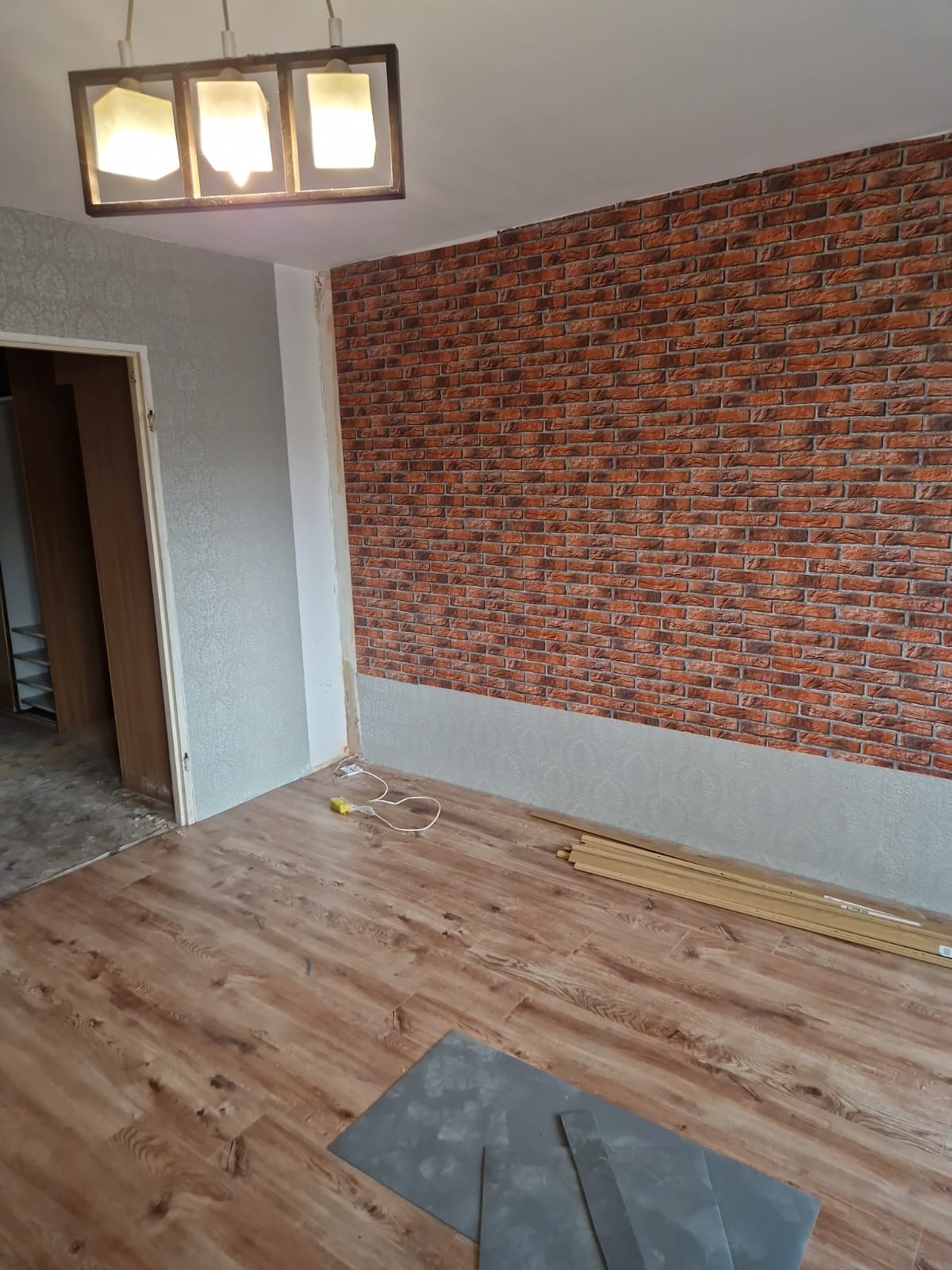 Pokój po częściowym remoncie: ułożone panele podłogowe, ściana z tapetą imitującą cegłę, widoczne listwy przypodłogowe i narzędzia.
