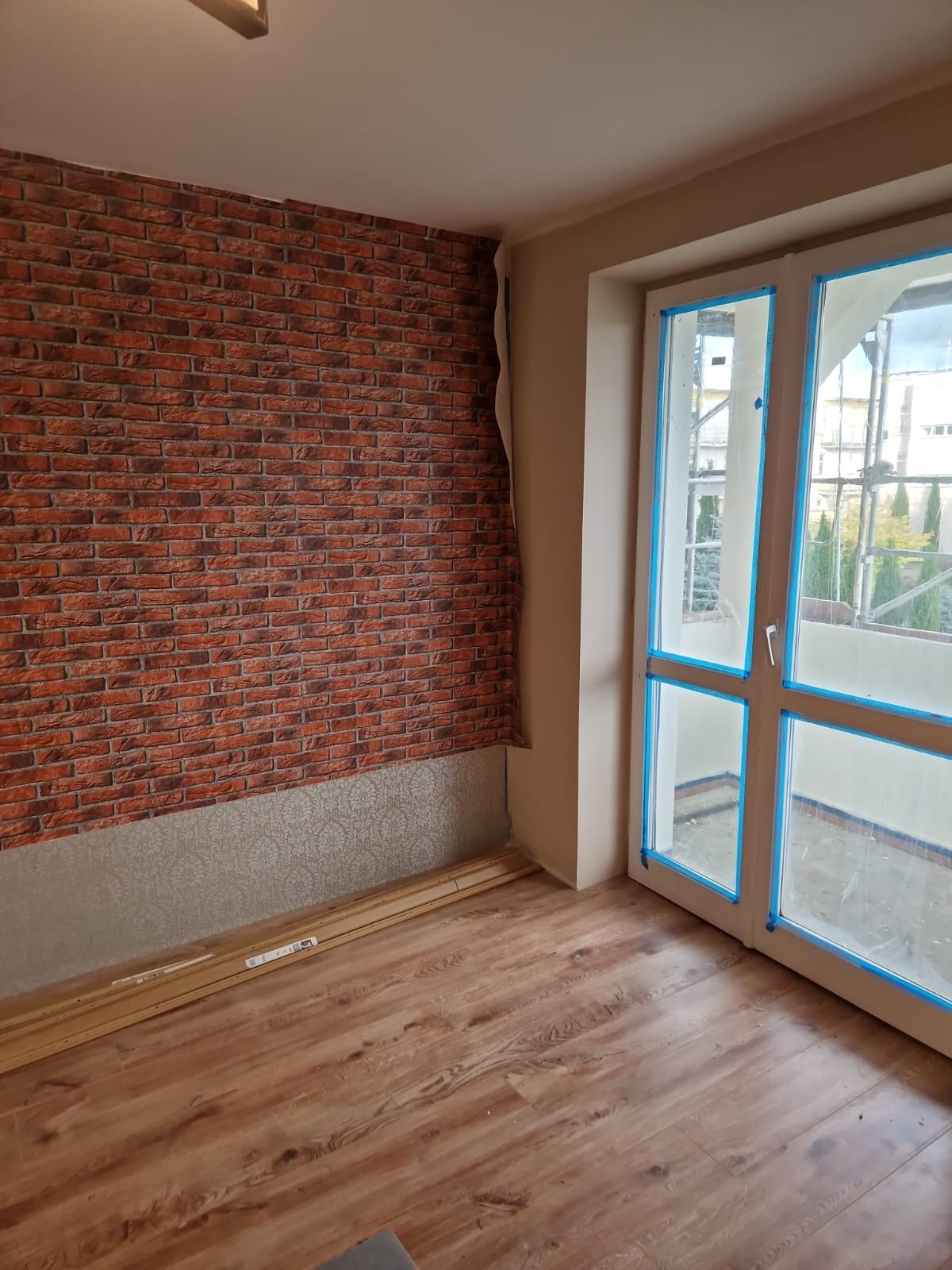 Pokój po remoncie: brązowe panele podłogowe, ściana z tapetą imitującą cegłę. Okno zabezpieczone folią malarską, listwy przypodłogowe do montażu.