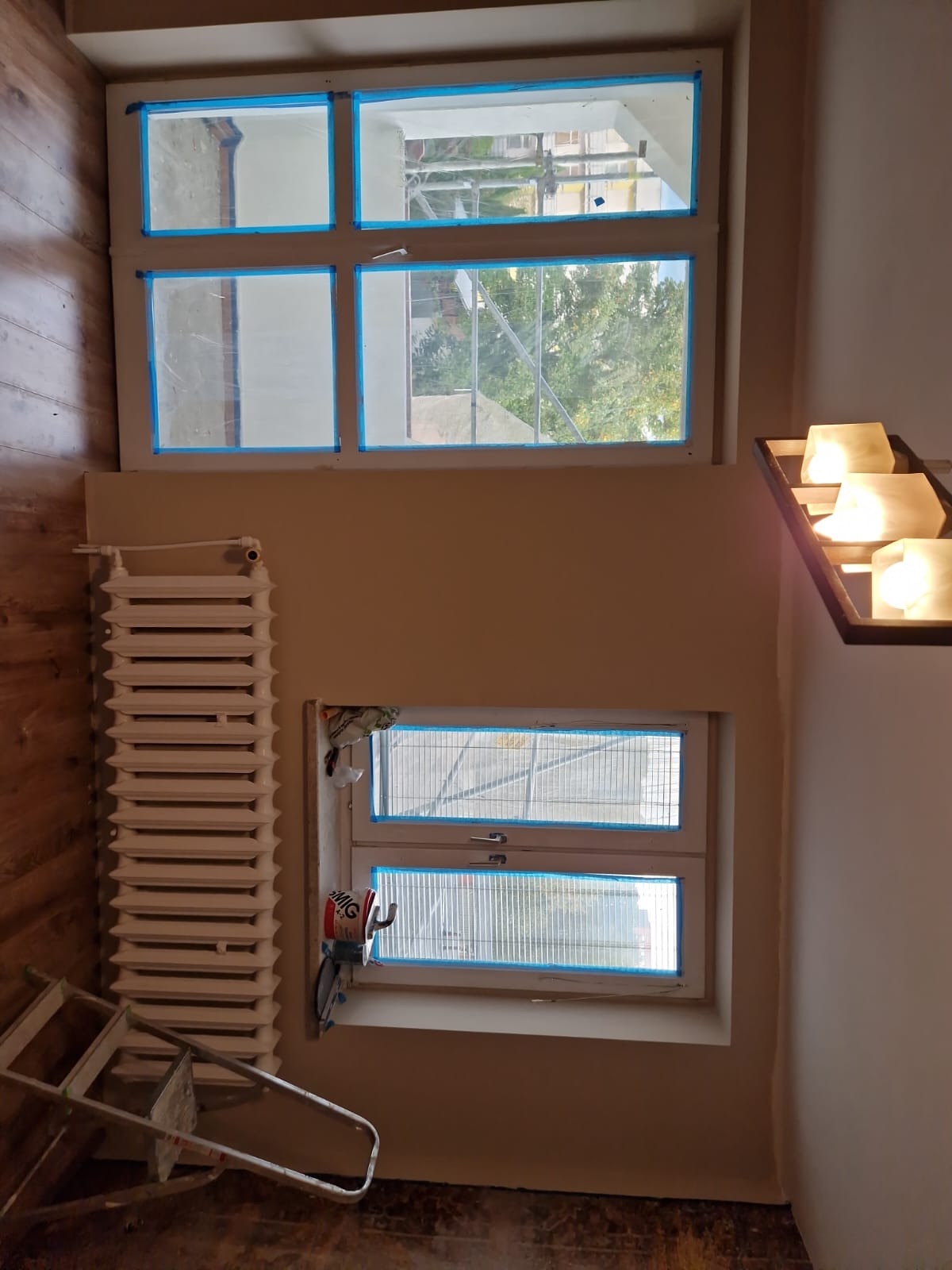 Przygotowanie do malowania: okna zabezpieczone taśmą malarską, grzejnik, drabina, widok na zewnątrz. Neutralne kolory ścian, drewniana podłoga.