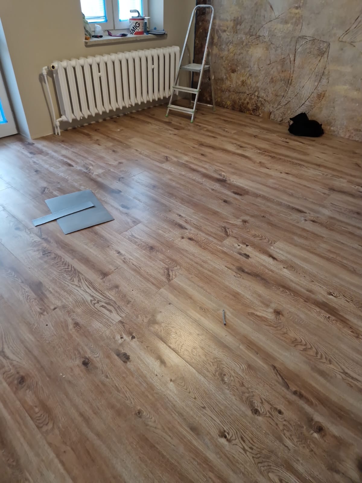Ukończona podłoga z paneli drewnopodobnych w pokoju z grzejnikiem i drabiną. Dwa panele leżące luzem na podłodze. Widoczne ślady po rysunku na ścianie.
