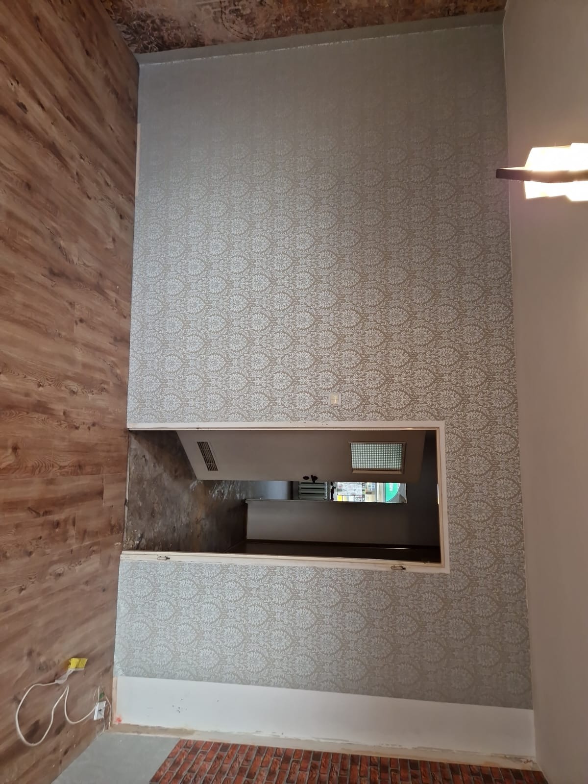 Wykończone ściany z wzorzystą tapetą i panele podłogowe, widok na otwarte przejście do innego pomieszczenia. Część sufitu z odsłoniętą strukturą.