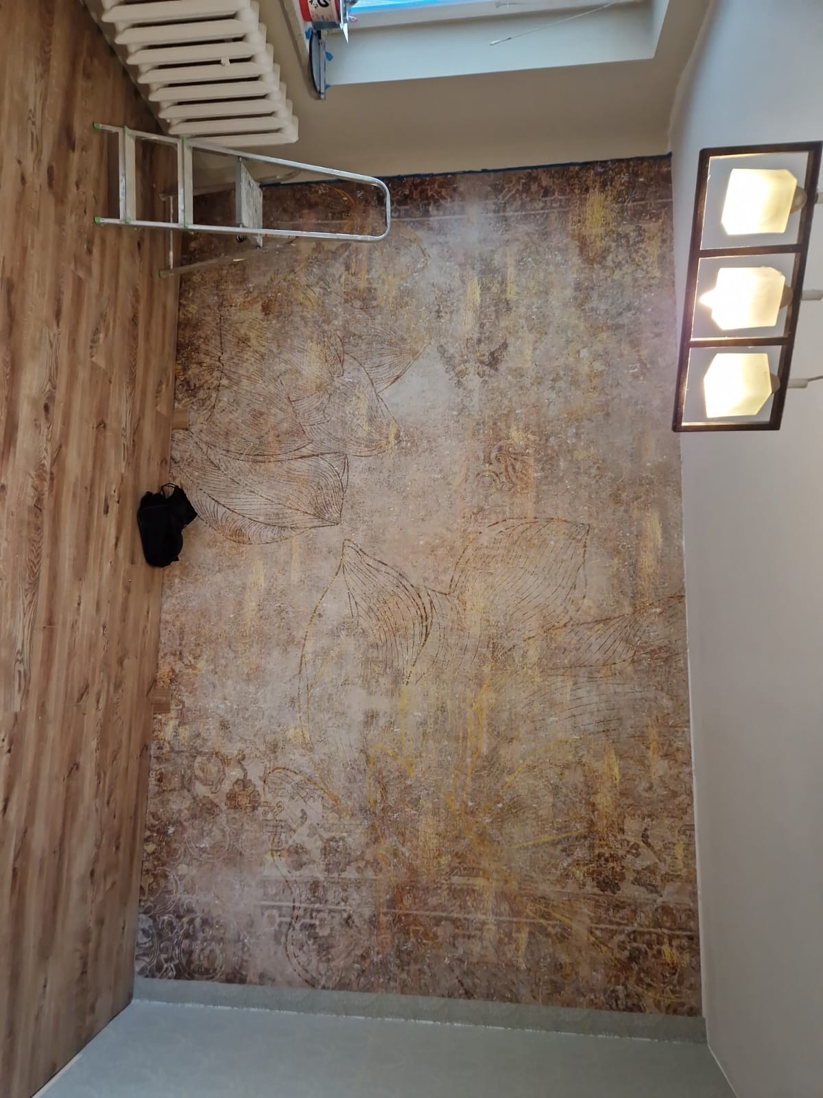 Wykończona ściana z dekoracyjnym wzorem liści w odcieniach brązu i złota, widoczna drabina, fragment grzejnika, oświetlenie sufitowe i drewnopodobna okładzina ściany.