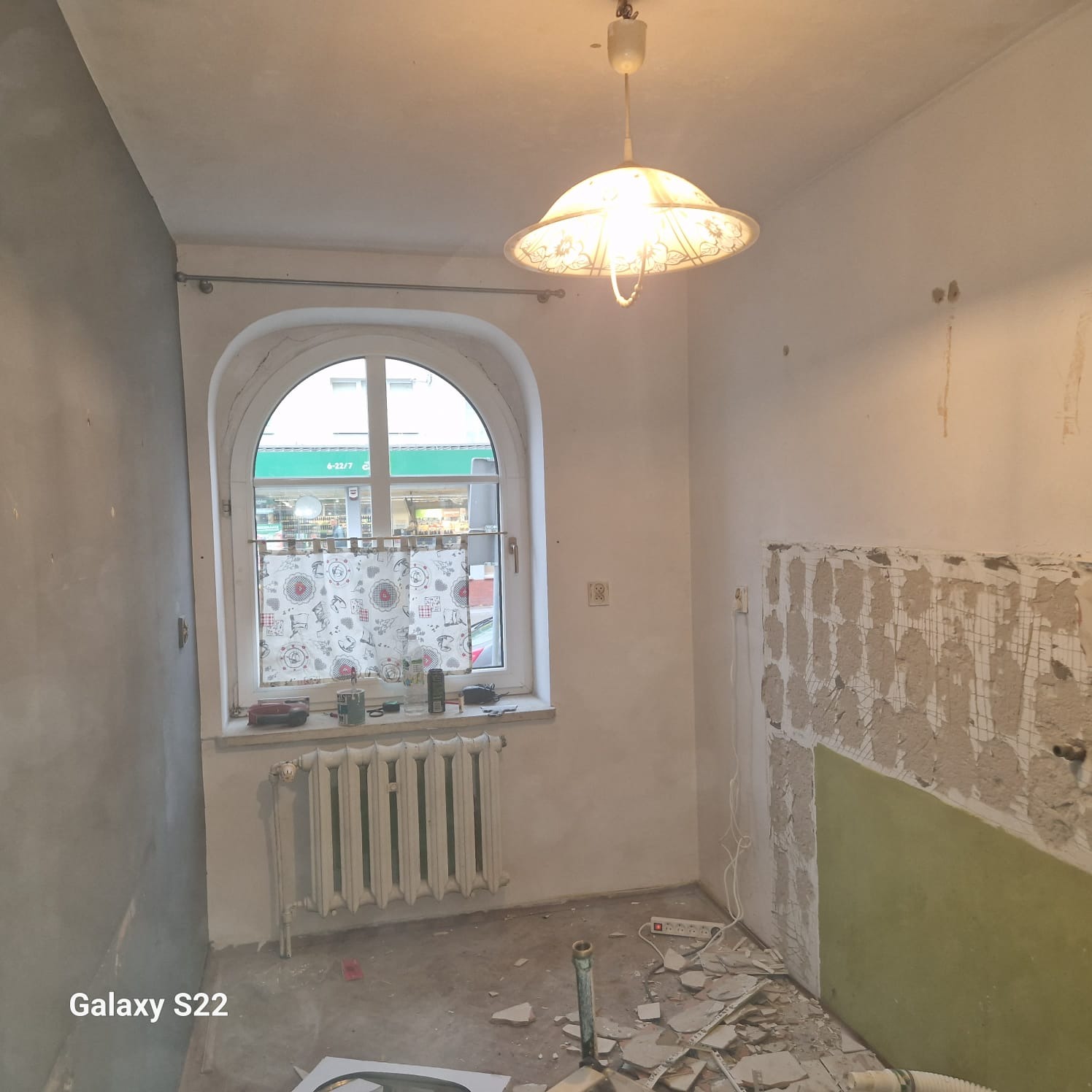 Fragment łazienki w trakcie demontażu płytek, z widocznym fragmentem ściany z siatką i resztkami kleju. Na podłodze gruz i elementy instalacji. Okno z zaokrąglonym łukiem.