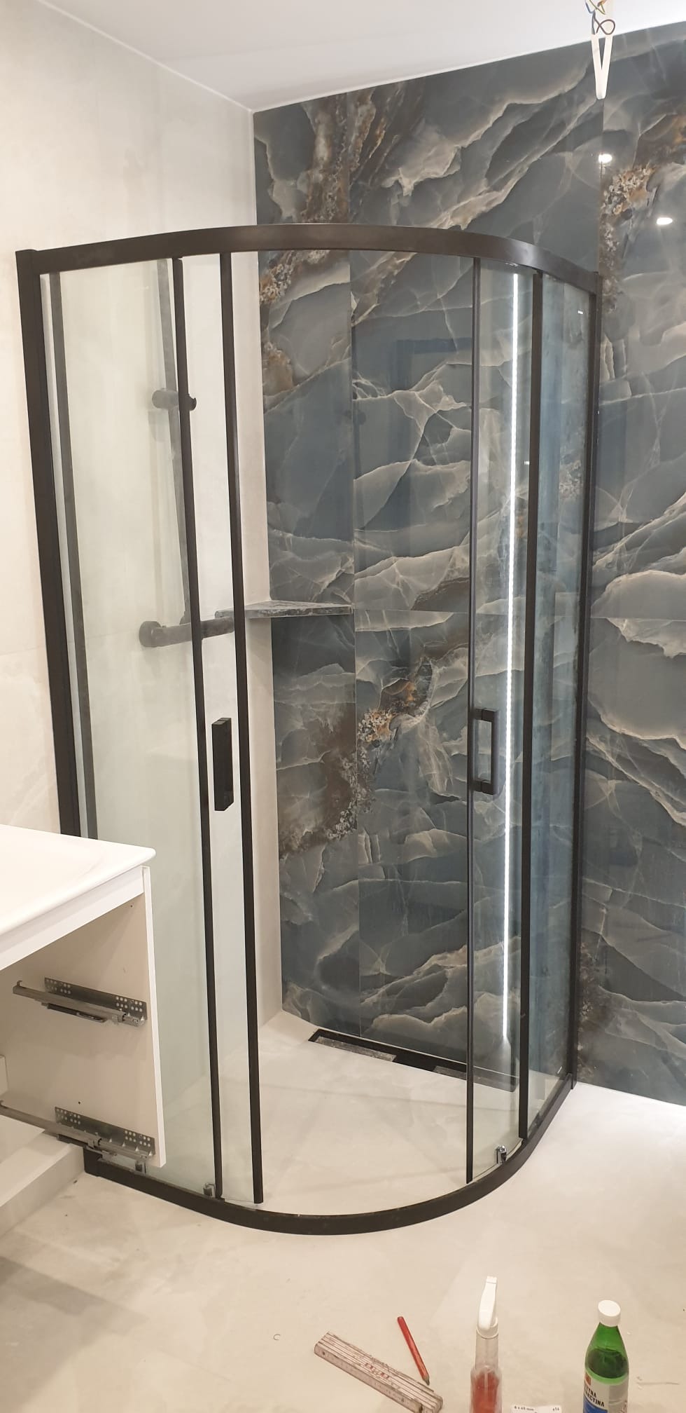 Narożna kabina prysznicowa z czarnymi profilami na tle ściany z płytek imitujących marmur. Widoczny fragment szafki umywalkowej i akcesoria na podłodze.