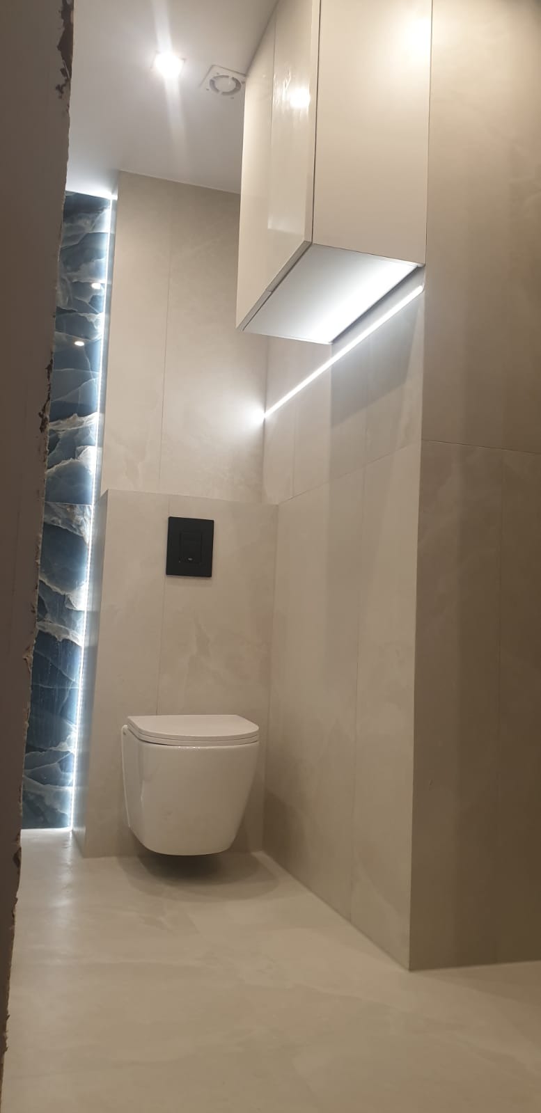 Nowoczesna łazienka z podwieszaną toaletą, szafką i oświetleniem LED. Beżowe płytki imitujące marmur, czarny przycisk spłuczki i niebieski akcent dekoracyjny.