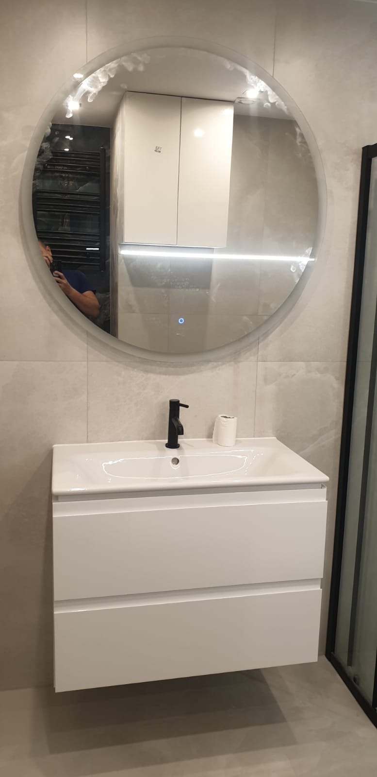 Biała szafka pod umywalkę z czarną armaturą i okrągłym lustrem z podświetleniem LED w nowoczesnej łazience z szarymi płytkami.