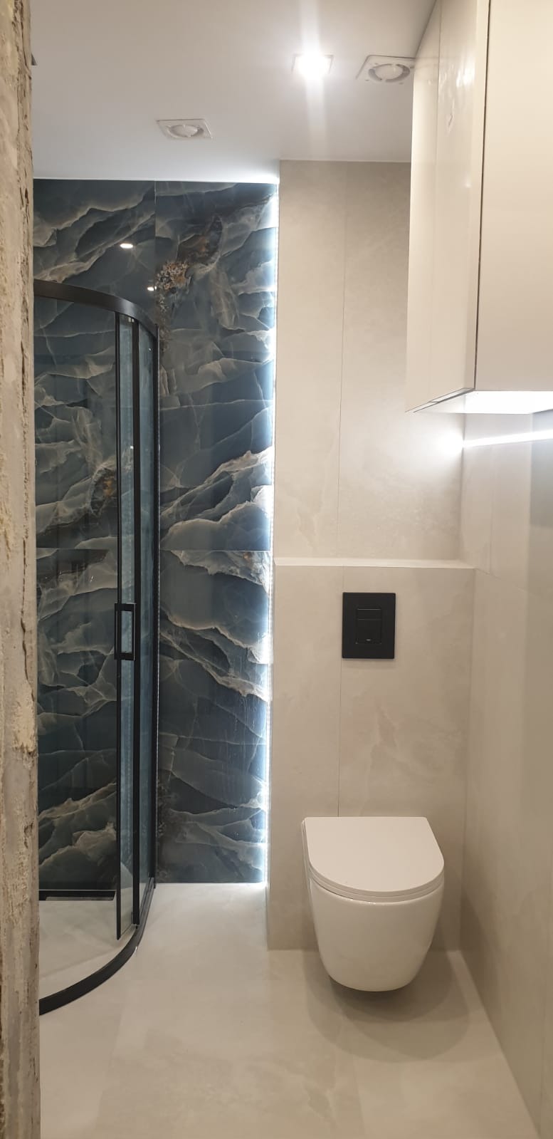 Nowoczesna łazienka z kabiną prysznicową z czarnymi ramami i płytkami imitującymi marmur, podświetlona listwą LED. Wisząca toaleta i szafka w jasnych odcieniach.