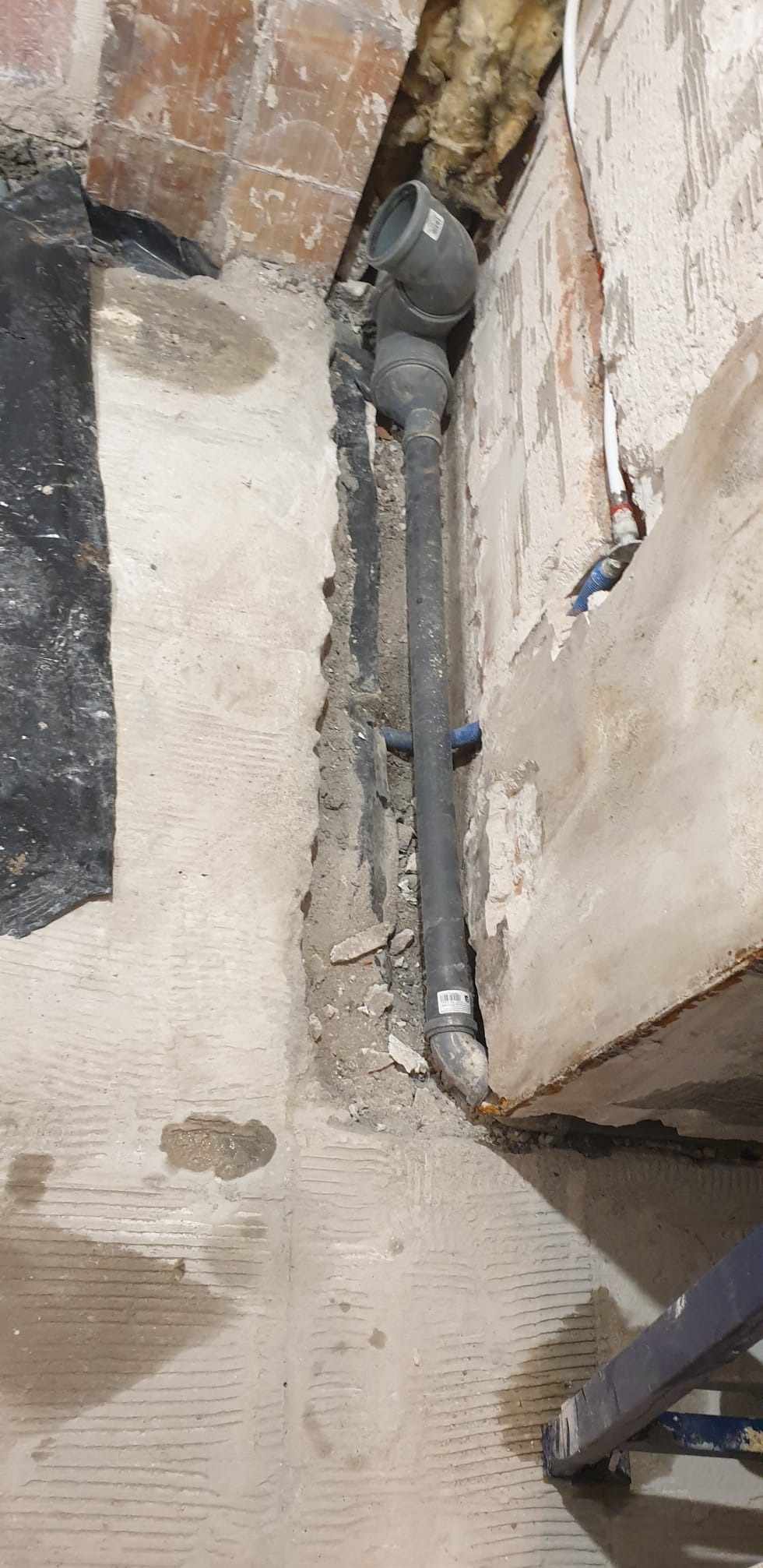 Szara rura kanalizacyjna w trakcie montażu w wykopie w ścianie, widoczne fragmenty instalacji wodnej i surowa ściana z cegły. Prace instalacyjne w łazience.