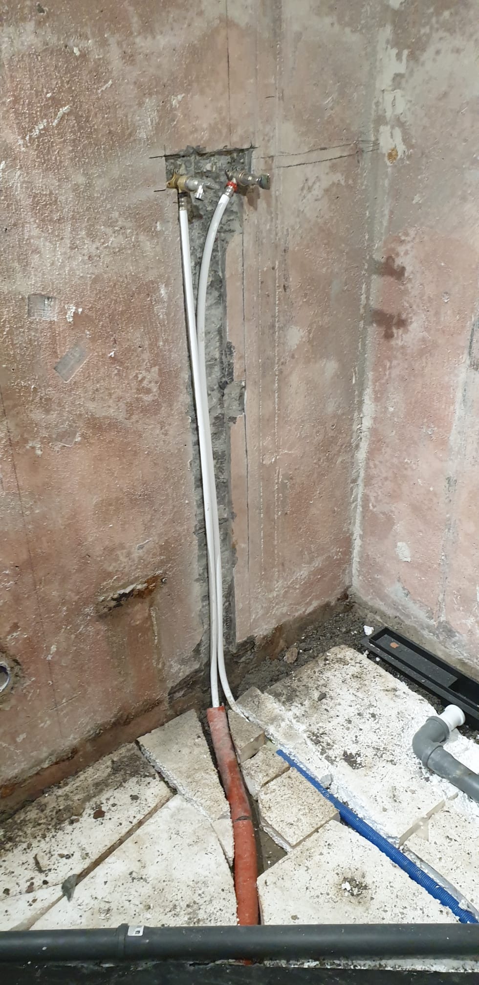 Ściana łazienki w trakcie remontu z widocznymi rurami instalacji wodnej i kanalizacyjnej, bruzdami w ścianie i fragmentami starej glazury na podłodze.