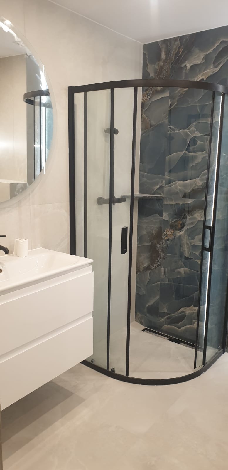 Nowoczesna łazienka z kabiną prysznicową typu walk-in z czarnymi profilami, białą szafką umywalkową i okrągłym lustrem. Ściana wykończona płytkami imitującymi marmur.