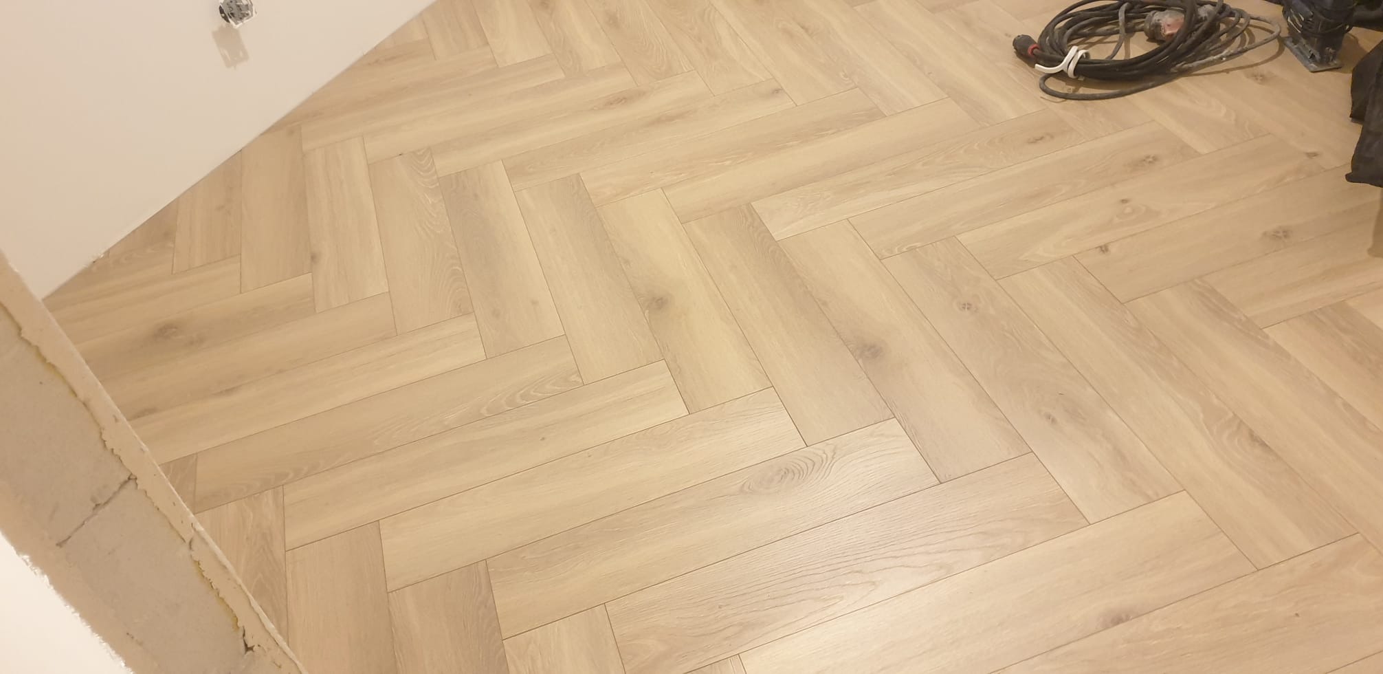 Jasne panele ułożone w jodełkę, widoczne narzędzia pracy w rogu pomieszczenia, ściana w trakcie wykończenia.