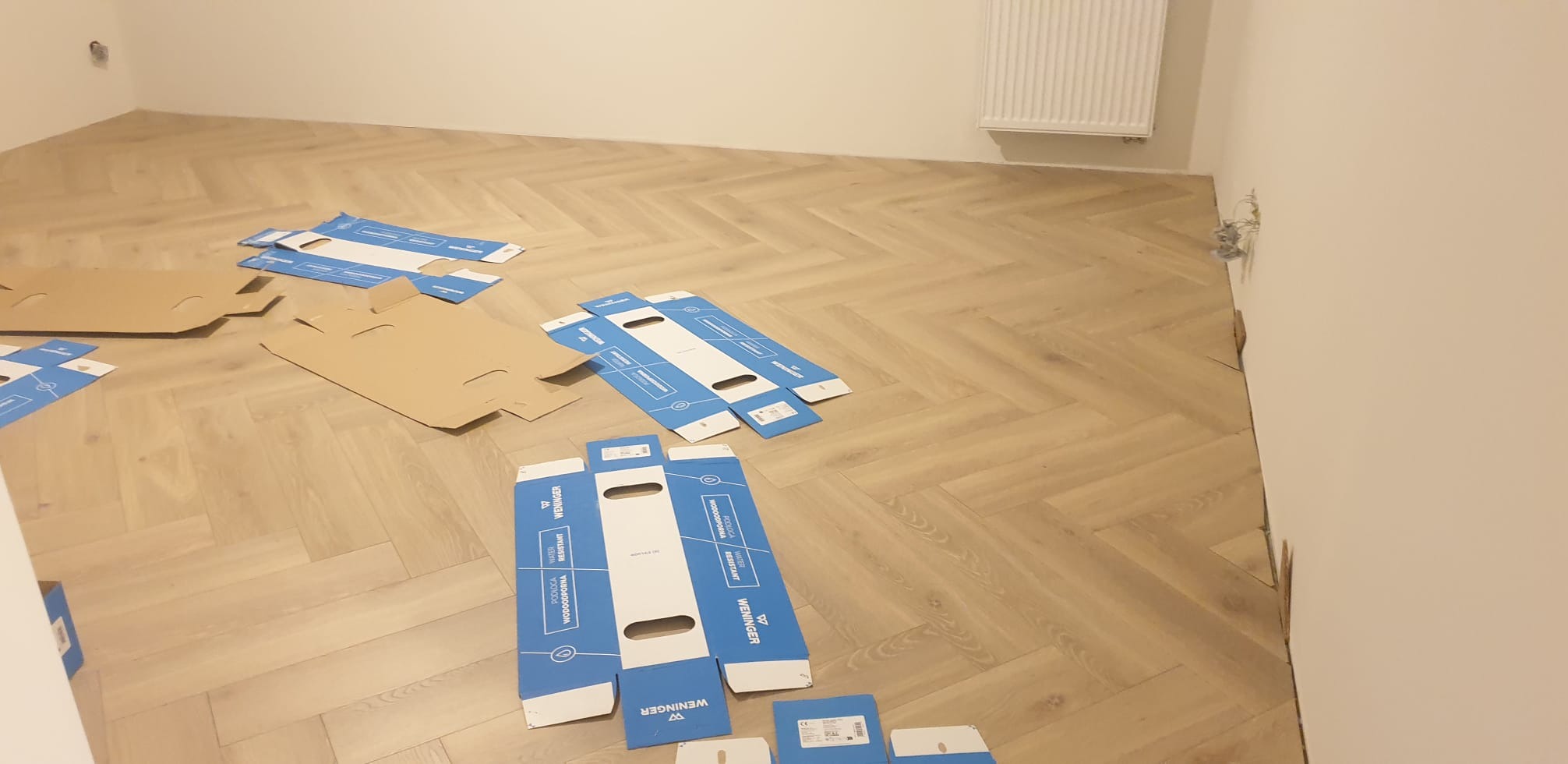 Ułożone panele podłogowe w jodełkę, z rozłożonymi kartonami po panelach. Widok na jasną podłogę i fragment ściany z grzejnikiem.