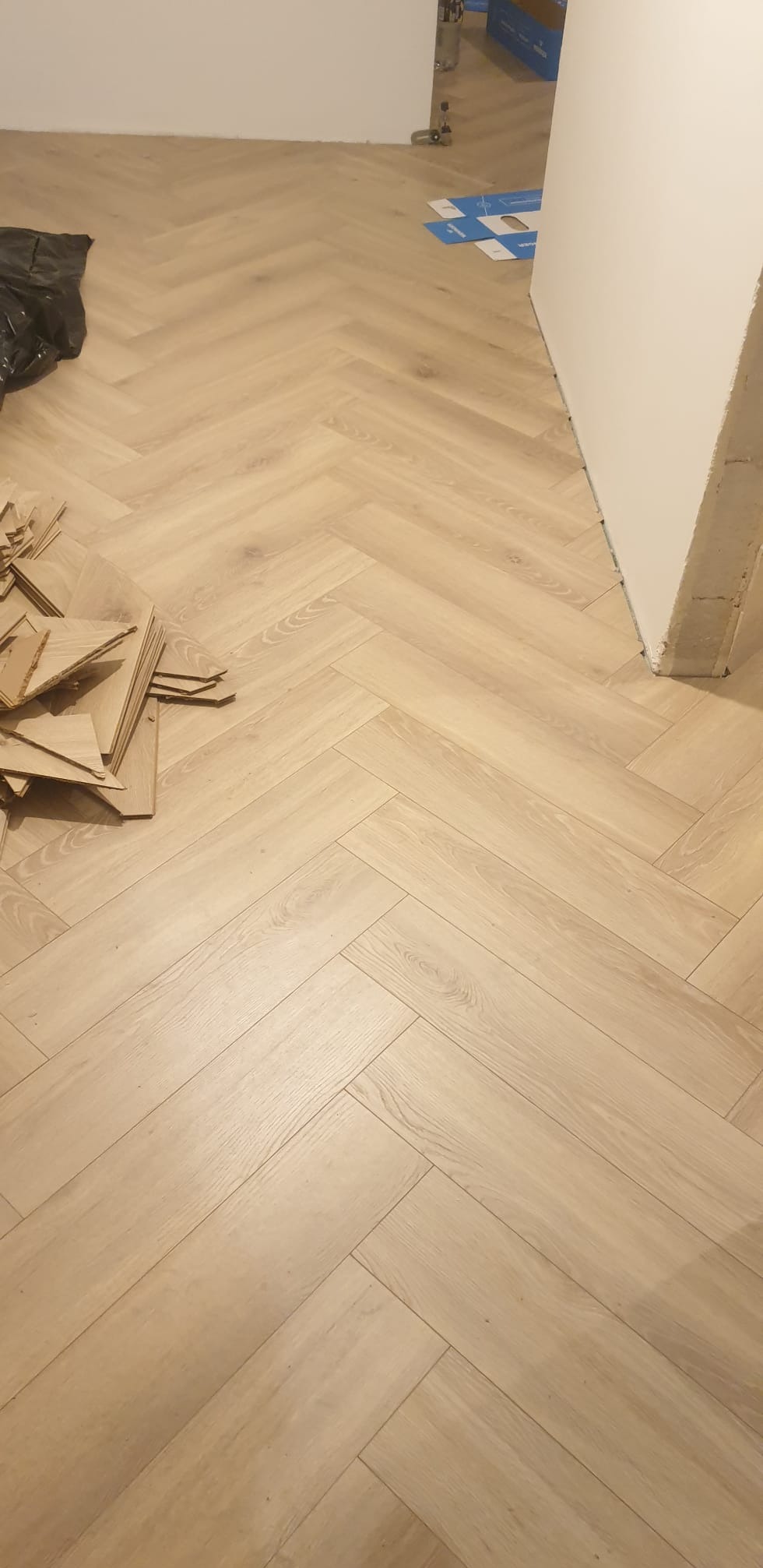 Jasne panele ułożone we wzór jodełki francuskiej, z widocznymi resztkami materiału i workiem, sugerujące etap prac wykończeniowych w pomieszczeniu.