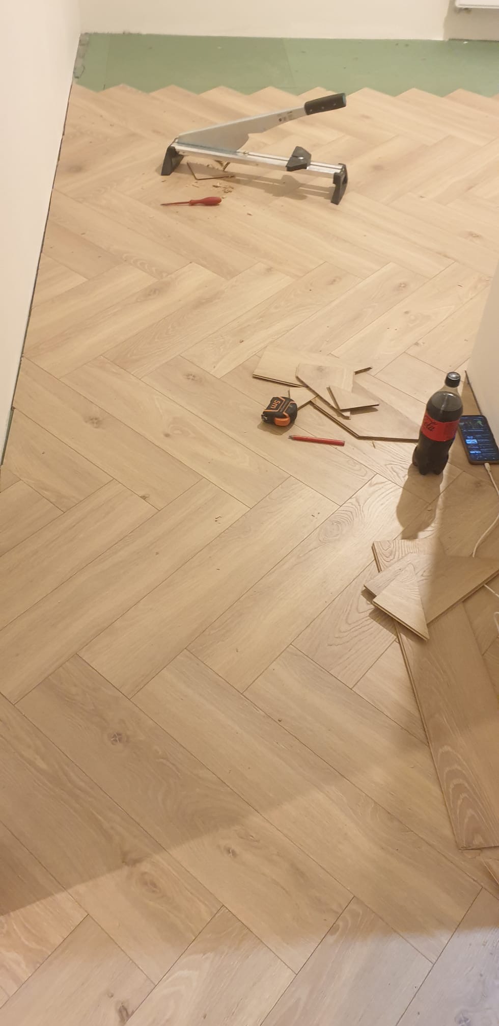 Układanie jasnych paneli podłogowych w jodełkę, widoczny nóż do paneli, miarka, ołówek i niedokończony fragment podłogi przy ścianie.