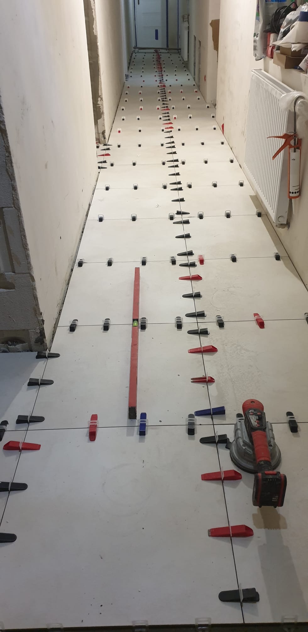 Białe płytki podłogowe w korytarzu, w trakcie układania, z użyciem poziomicy i klipsów dystansowych. W tle widoczny grzejnik i narzędzia glazurnicze.