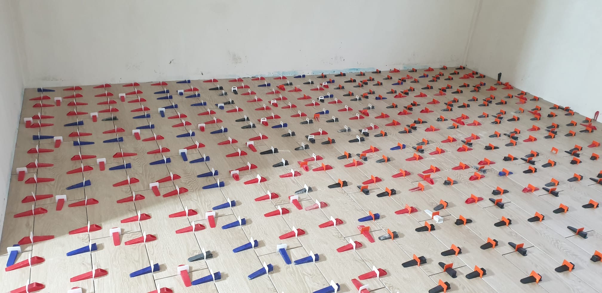 Układanie płytek podłogowych z użyciem systemu poziomowania, widoczne kliny i klipsy w kolorach czerwonym, niebieskim, pomarańczowym i czarnym na jasnych płytkach.