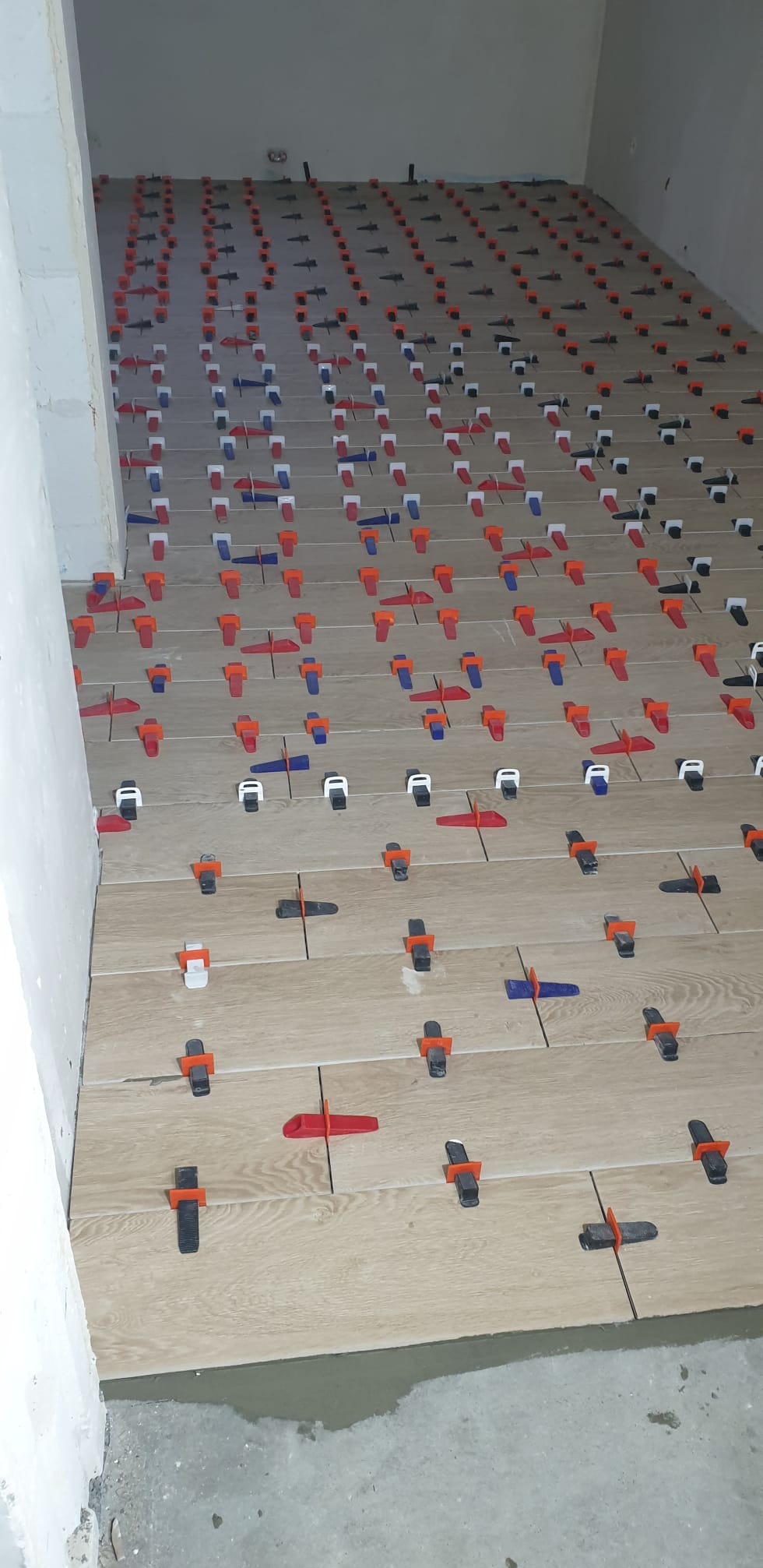 Układanie płytek podłogowych z użyciem systemu poziomowania; widoczne klipsy i kliny w kolorach czerwonym, niebieskim, białym i czarnym. Jasne płytki imitujące drewno.