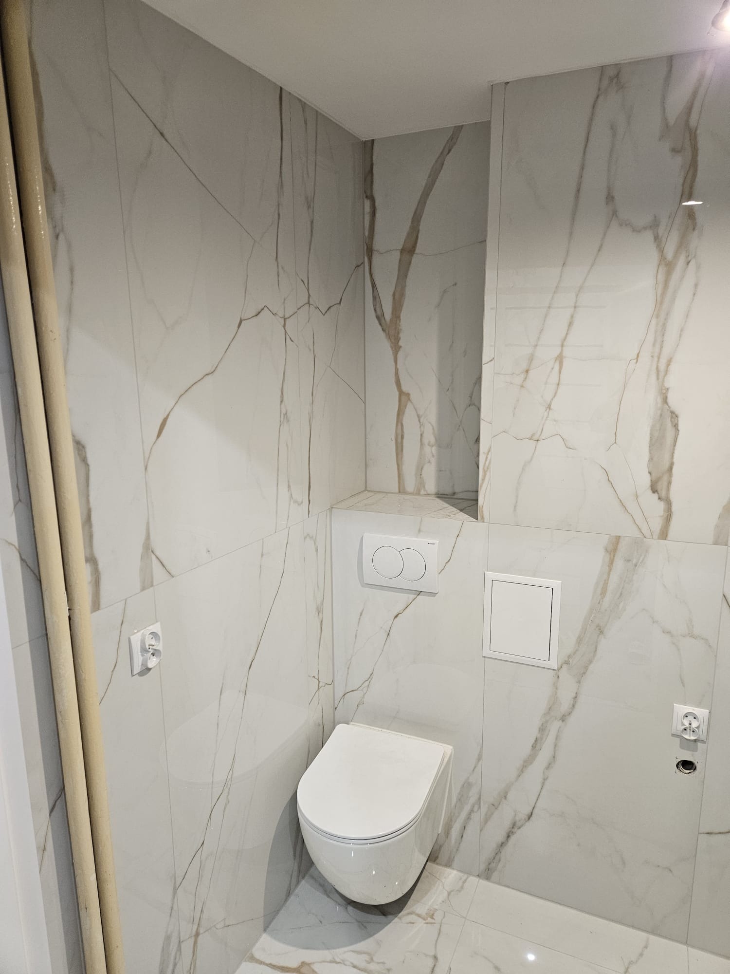 Biała, marmurkowa łazienka z podwieszaną toaletą, widoczne rury i gniazdka elektryczne. Minimalistyczny design z akcentem na naturalny wzór kamienia.